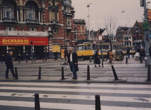 A yellow and grey articulated tram passes a red-brick theatre with a red Richard III banner while a person in a dark coat poses beside bollards next to a zebra crossing.