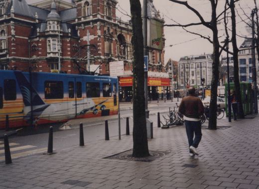 A blue, orange and yellow tram painted with an airliner runs along a street in front of an ornate red-brick theatre with red Richard III banners, while a pedestrian walks away from the camera along a wet brick sidewalk lined with bare trees and bollards.