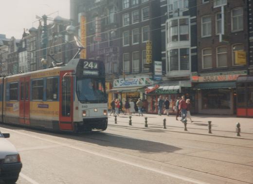 An articulated tram with a grey front and red doors, wrapped in yellow and blue advertising and showing route 24 Stationsplein, moves along a city street in front of shops and pedestrians, with bollards along the sidewalk.