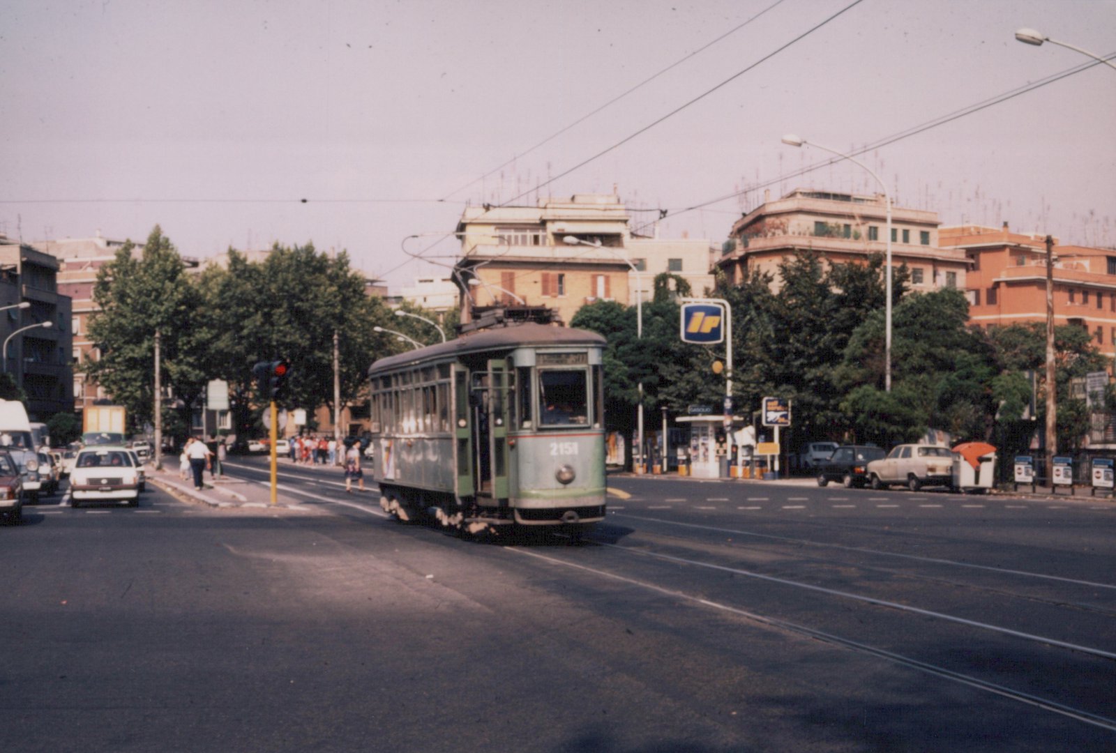 A green single-unit streetcar with a pantograph runs on street track toward the camera on a wide boulevard, with apartment buildings, trees, a filling station and a red traffic light.