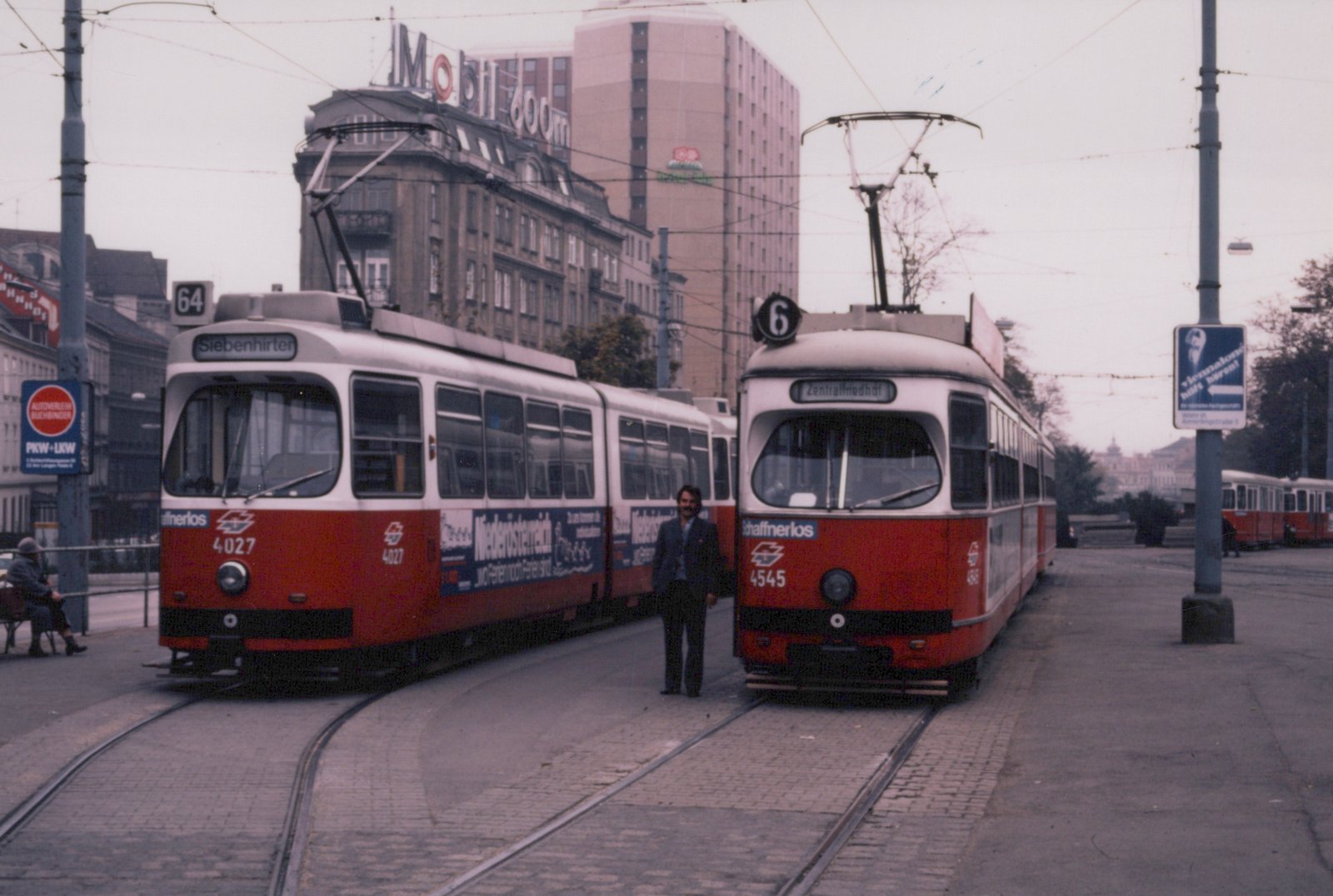 Two red and white articulated trams face the camera on cobblestone tracks, with a man standing between them and city buildings and a tall hotel tower behind.