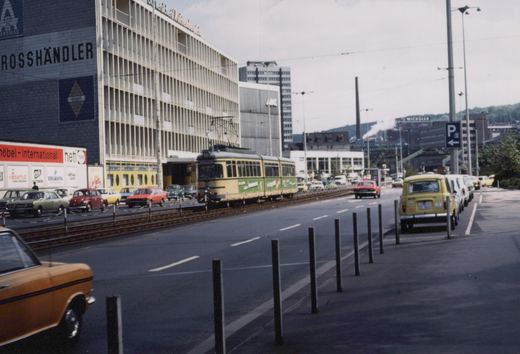 A cream and green three-section tram with a pantograph on a fenced track down the middle of a wide street, with modern office buildings and parked cars at left, the Wicküler brewery sign on a hill in the distance, and a line of bollards in the foreground.
