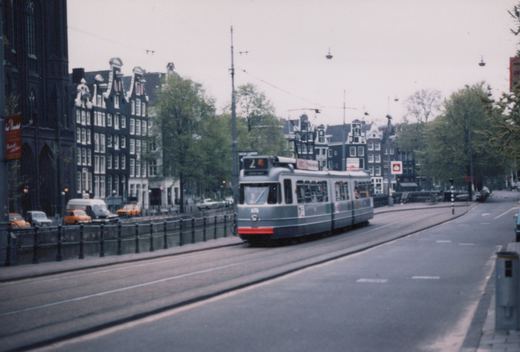 A grey three-section articulated tram with a red front bumper and route number 2 runs on tracks beside a railed canal, with gabled canal houses, trees and a dark church behind it.