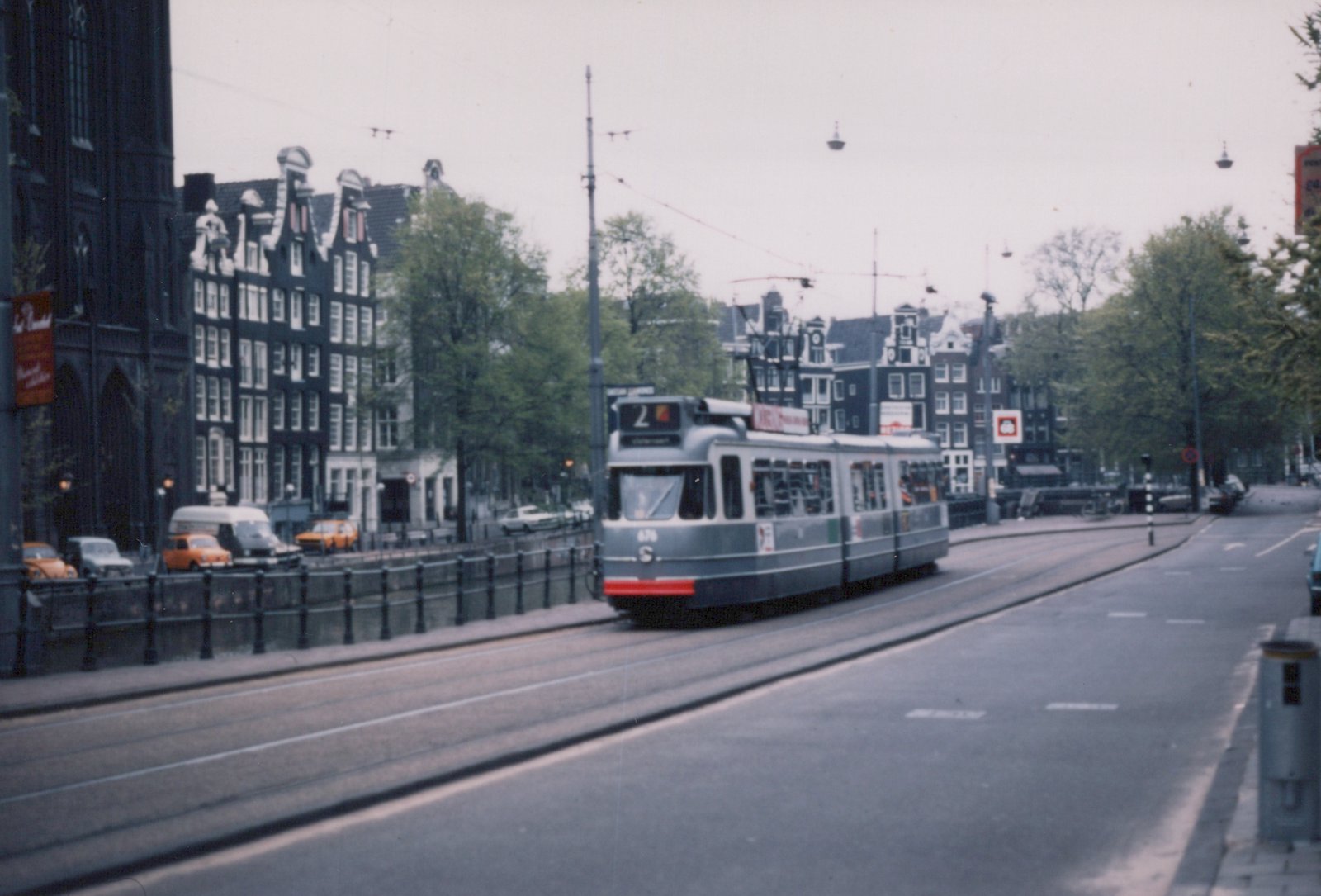 A grey three-section articulated tram with a red front bumper and route number 2 runs on tracks beside a railed canal, with gabled canal houses, trees and a dark church behind it.