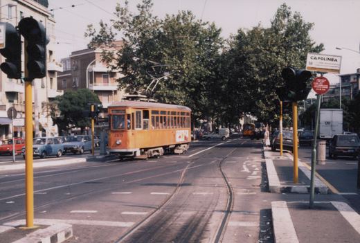 An orange single-ended streetcar with a pantograph rounds a curve on double track in a tree-lined avenue, with traffic lights, parked cars and a CAPOLINEA sign in the foreground.