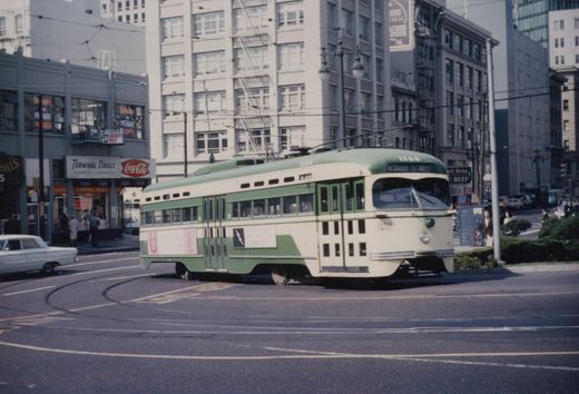 A green and cream streetcar numbered 1144 turns on curving street track beneath overhead wires, with the Terminal Drugs store and downtown office buildings behind it.
