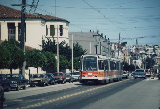 A white light rail train with a red and orange band, made up of two articulated cars, comes down a sunny street with rails in the pavement. Cars are parked along the left curb in front of a white stucco building with a tile roof, and hillside houses and a church spire are in the distance.