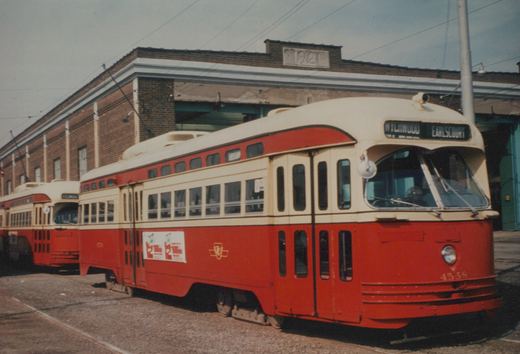 A red and cream streetcar numbered 4558 parked on a cobbled yard in front of a brick carhouse with a 1921 date stone, with a second similar streetcar behind it.