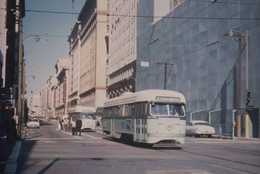 A green and white streetcar turns through an intersection on a sunny downtown street beneath overhead wires, with a second streetcar, pedestrians and tall buildings behind it.
