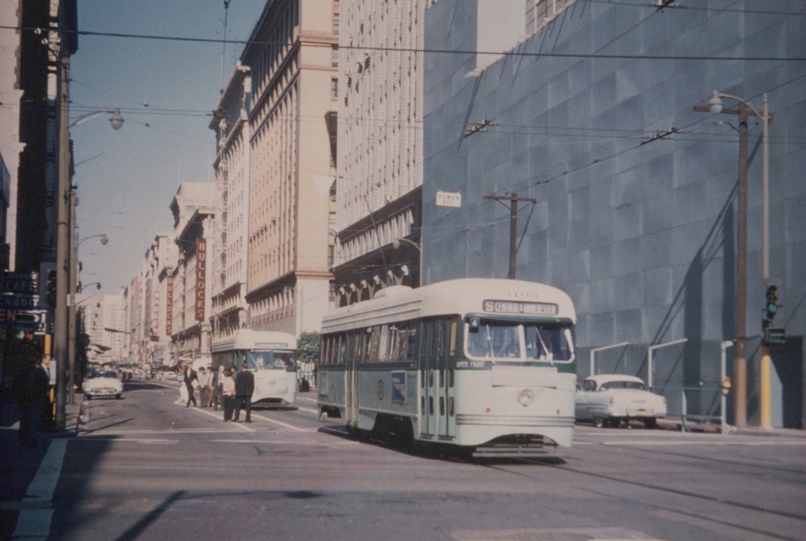 A green and white streetcar turns through an intersection on a sunny downtown street beneath overhead wires, with a second streetcar, pedestrians and tall buildings behind it.