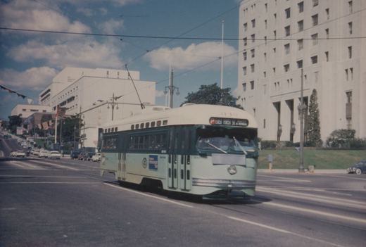 A green and cream streetcar with a trolley pole runs along a wide street under overhead wires, with tall white buildings and parked cars behind it.