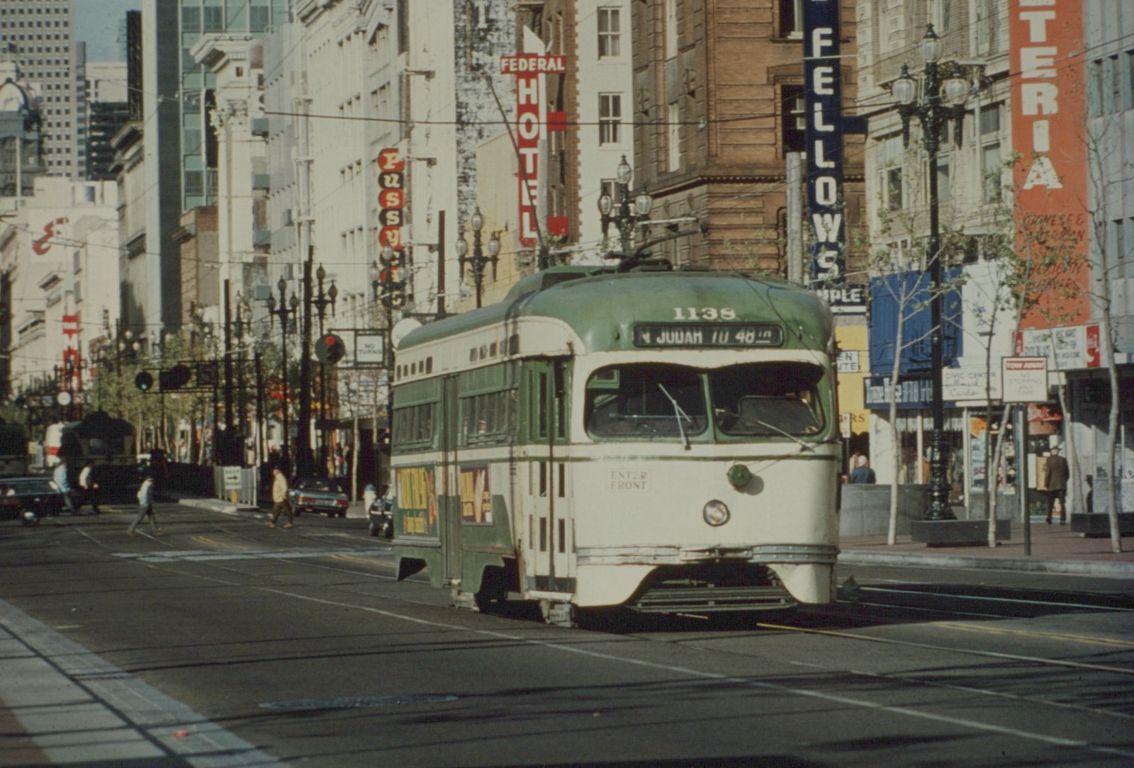 A green and cream streetcar with destination sign N JUDAH TO 48th runs along a city street lined with older buildings and vertical hotel and shop signs while pedestrians cross behind it.