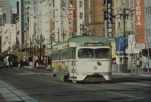 A green and cream streetcar with destination sign N JUDAH TO 48th runs along a city street lined with older buildings and vertical hotel and shop signs while pedestrians cross behind it.