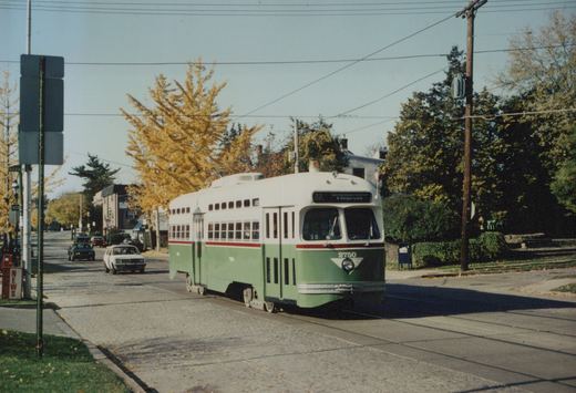 A green and cream streetcar numbered 2750, signed 23 Germantown Avenue & Gorgas Lane, travels on a cobbled street under overhead wires, with yellow autumn trees and cars behind it.