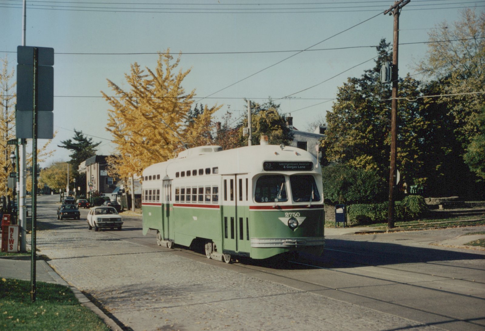 A green and cream streetcar numbered 2750, signed 23 Germantown Avenue & Gorgas Lane, travels on a cobbled street under overhead wires, with yellow autumn trees and cars behind it.