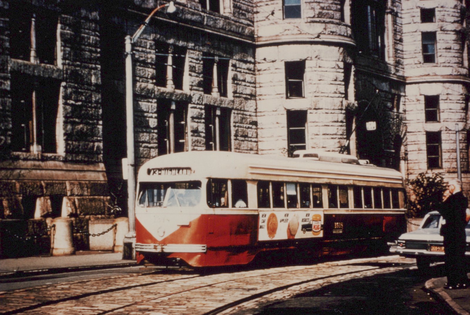 A red and cream streetcar with a 73-HIGHLAND sign rounds a curve on cobblestone tracks in front of a dark stone building with round towers, while a man stands beside a parked white car at right.