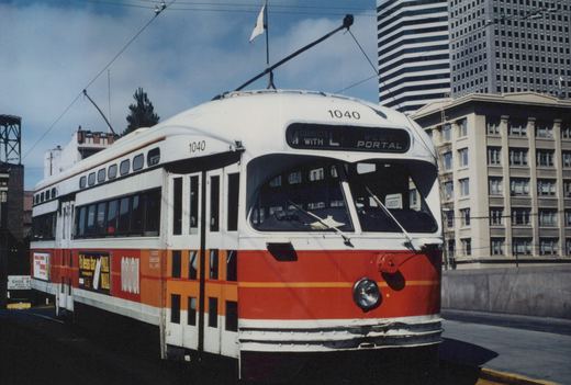 An orange, red and white streetcar with its trolley pole raised and a sign reading M connects with L West Portal stands on a terminal ramp, with downtown high-rise buildings behind it.