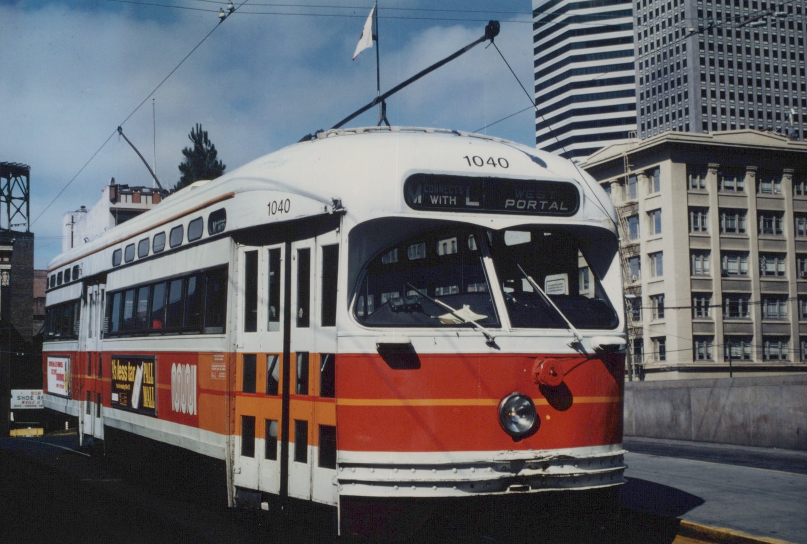 An orange, red and white streetcar with its trolley pole raised and a sign reading M connects with L West Portal stands on a terminal ramp, with downtown high-rise buildings behind it.