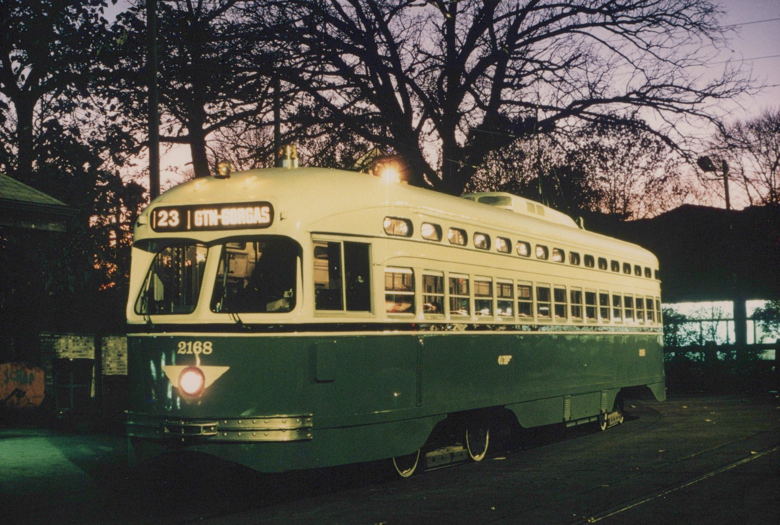 A green and cream streetcar with lit windows and headlight, numbered 2168, stands on track under bare trees against a purple dusk sky.