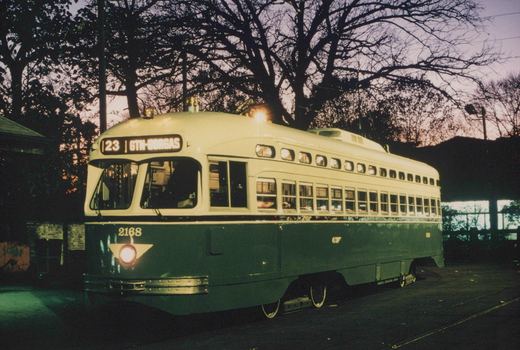 A green and cream streetcar with lit windows and headlight, numbered 2168, stands on track under bare trees against a purple dusk sky.