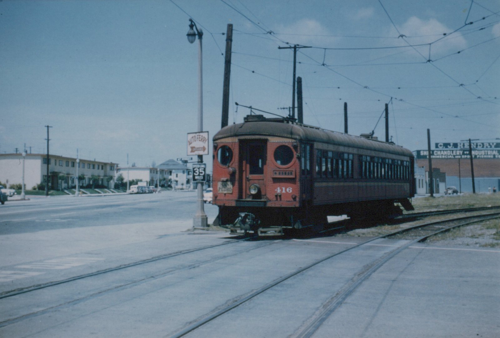 A red wooden interurban car with round porthole windows on its front end, numbered 416 and carrying a San Pedro sign, sits at a street crossing beneath a web of trolley wires, with apartment buildings and a ship chandlery building behind it.