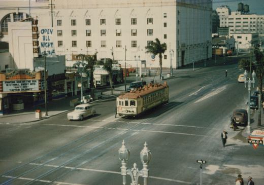 High view of a green and cream streetcar on double track at a broad intersection, with a theater marquee, shops, palm trees, a large white building and a few pedestrians and parked cars.