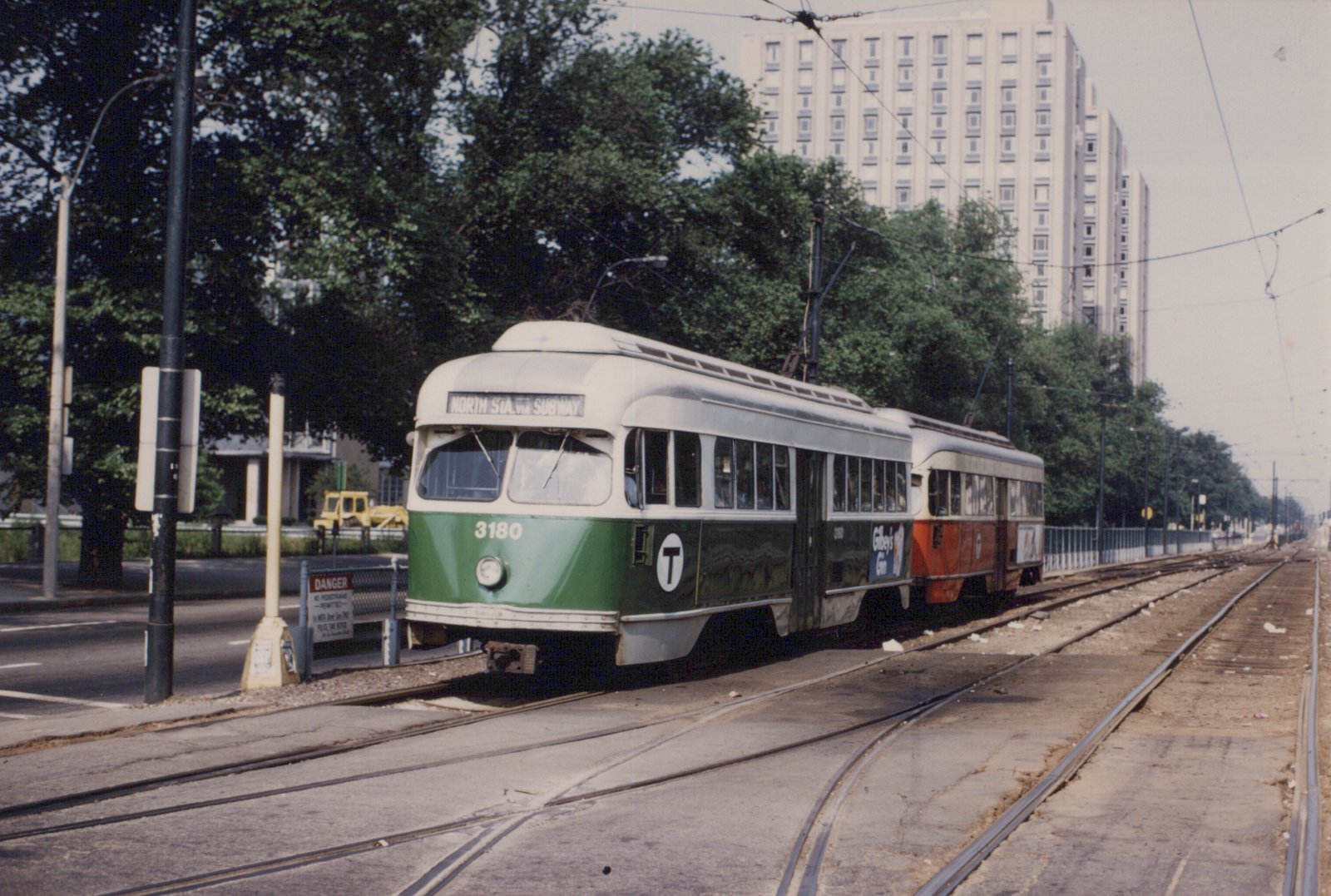 A green and cream streetcar numbered 3180 coupled to an orange and cream streetcar runs on paved double track in the median of a tree-lined avenue, with a tall apartment block behind.