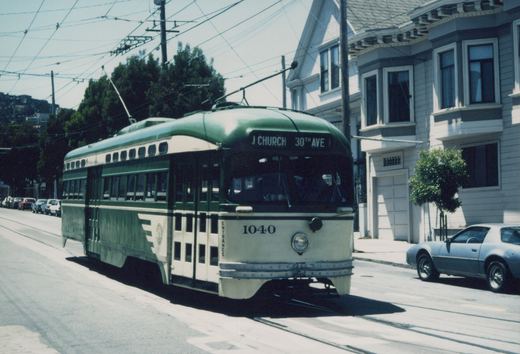 A green and cream streetcar numbered 1040 with a J CHURCH 30TH AVE sign runs on street track under overhead wires, past Victorian houses and a parked silver coupe.