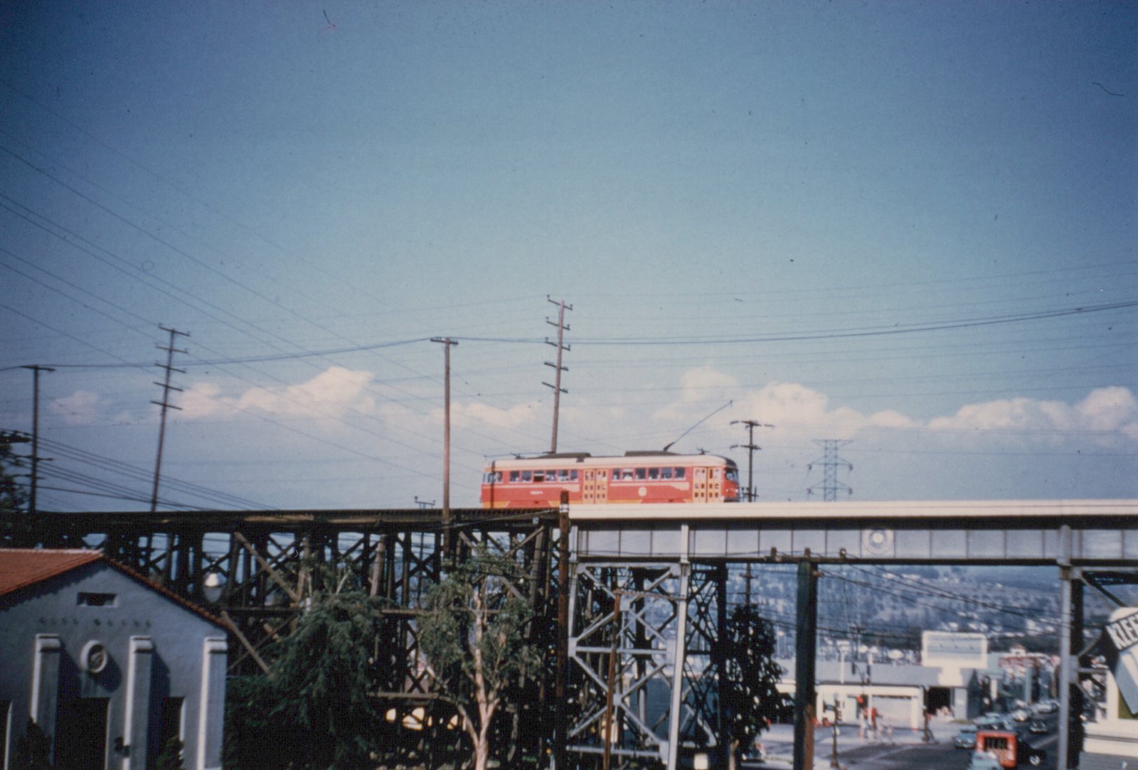 A red streetcar with a trolley pole crosses a long trestle with timber and steel spans against a blue sky. Utility poles and wires stand above it, a stucco building is at lower left, and a street with cars is at lower right.