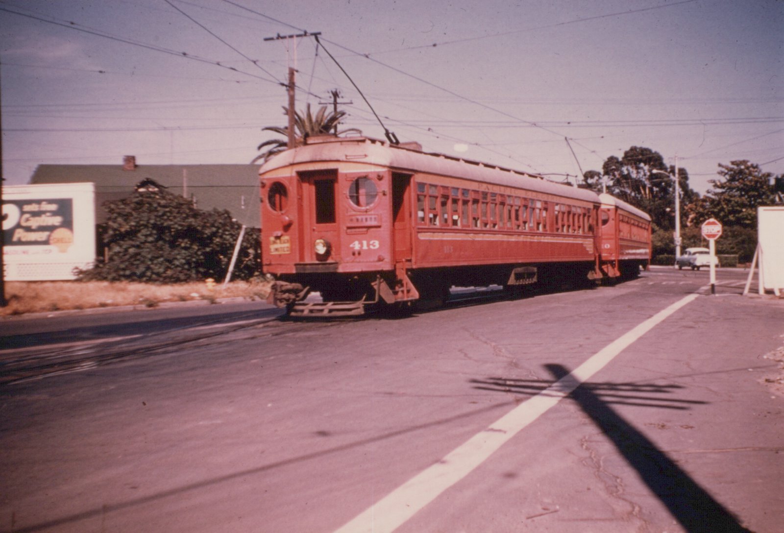 A two-car red interurban train with round end windows and trolley poles crosses a paved street under overhead wires, with a Shell billboard at left and a stop sign at right.