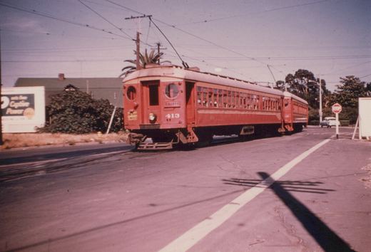 A two-car red interurban train with round end windows and trolley poles crosses a paved street under overhead wires, with a Shell billboard at left and a stop sign at right.