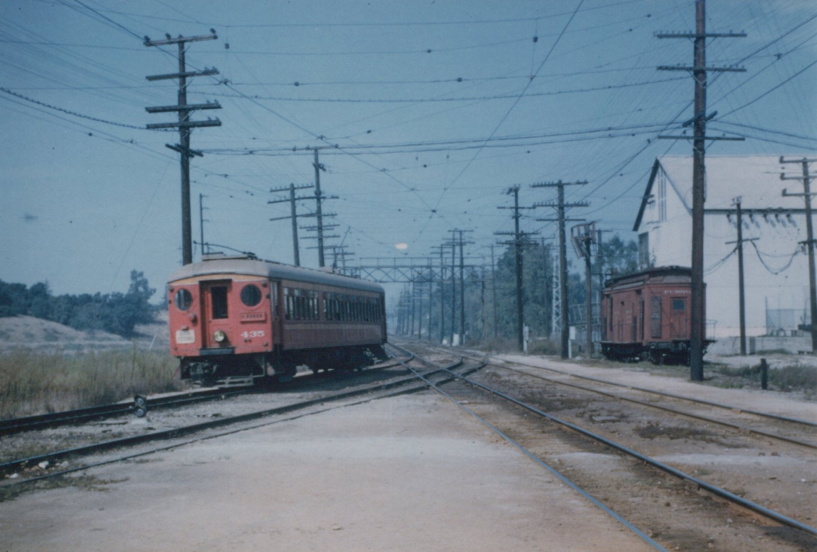 A red wooden interurban car with round end windows and a trolley pole rounds a curve at a multi-track junction under a dense web of overhead wires, with a red wooden car parked beside a white building at right.