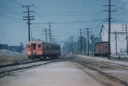 A red wooden interurban car with round end windows and a trolley pole rounds a curve at a multi-track junction under a dense web of overhead wires, with a red wooden car parked beside a white building at right.