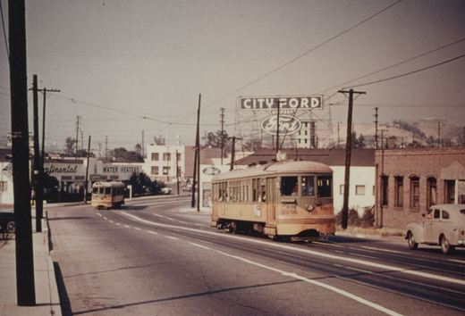 A yellow and green streetcar numbered 1387 with a W roof sign travels on a wide paved street under overhead wires, with a second streetcar behind it, a large City Ford sign on a tower, low buildings, a 1940s sedan and hills in the distance.