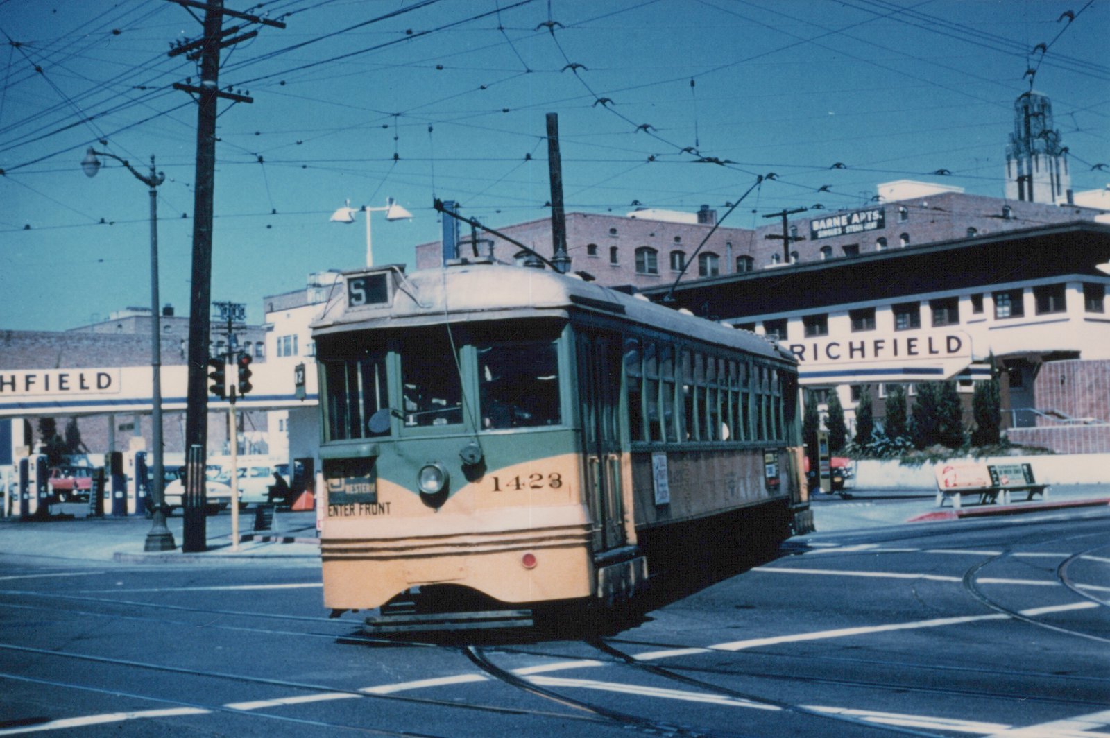 A green and yellow double-ended streetcar numbered 1423, showing S on its roof sign, curves through a street intersection under a web of overhead wires, with Richfield gas stations and brick apartment buildings behind it.