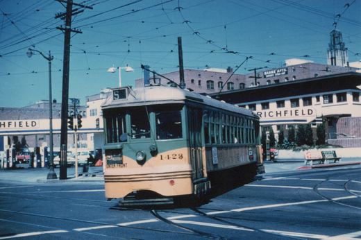 A green and yellow double-ended streetcar numbered 1423, showing S on its roof sign, curves through a street intersection under a web of overhead wires, with Richfield gas stations and brick apartment buildings behind it.