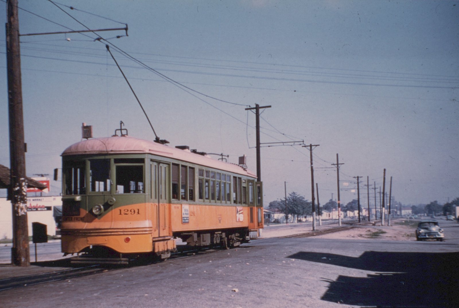 A yellow and green streetcar with its trolley pole raised stands on track beside a wide road lined with wooden utility poles and overhead wires, with a sedan parked at right and low buildings in the distance.
