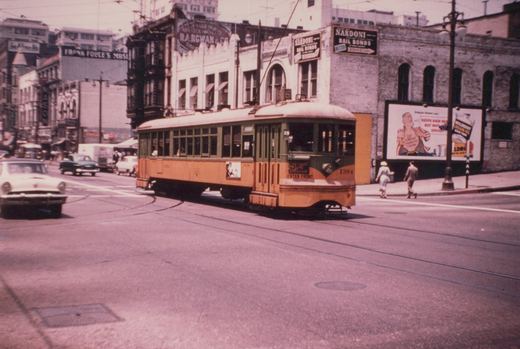 A yellow and green streetcar numbered 1394 crosses an intersection on curved track under trolley wire, while two pedestrians cross the street beside brick buildings with bail bond signs and a hair-tonic billboard.