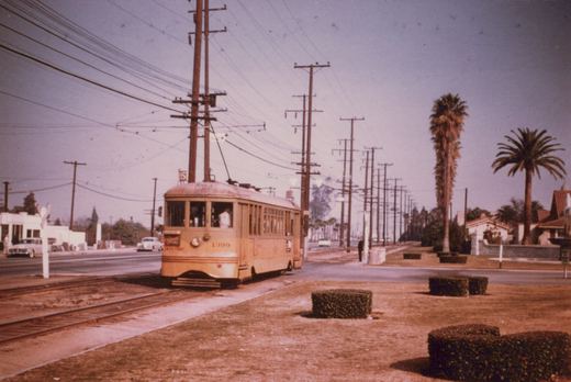A yellow streetcar numbered 1399 runs toward the camera on double track in a dirt median, with tall utility poles, palm trees and trimmed hedges along a wide boulevard.