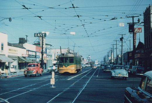A green and yellow single-ended streetcar with a trolley pole comes down a wide street with crossing tracks, a dense web of overhead wire, storefronts, and 1950s cars and a red pickup truck.