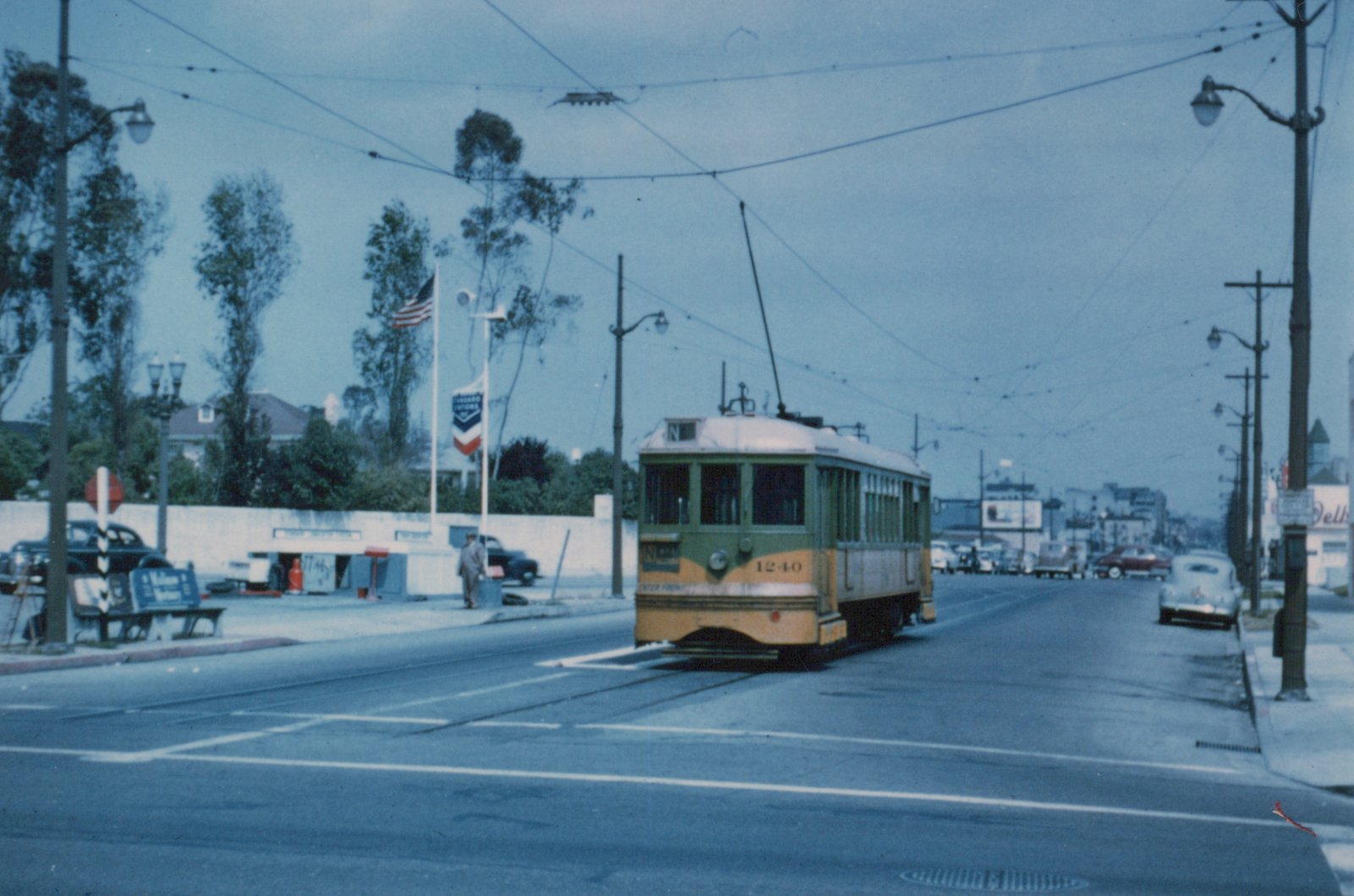 A yellow and green streetcar with a trolley pole and the number 1240 on its dash approaches a crosswalk on a wide street under overhead wires, with a gas station and a parked car on either side.