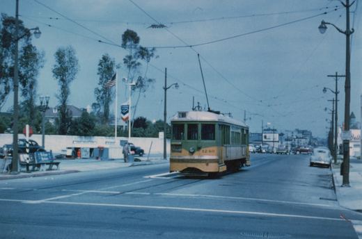 A yellow and green streetcar with a trolley pole and the number 1240 on its dash approaches a crosswalk on a wide street under overhead wires, with a gas station and a parked car on either side.