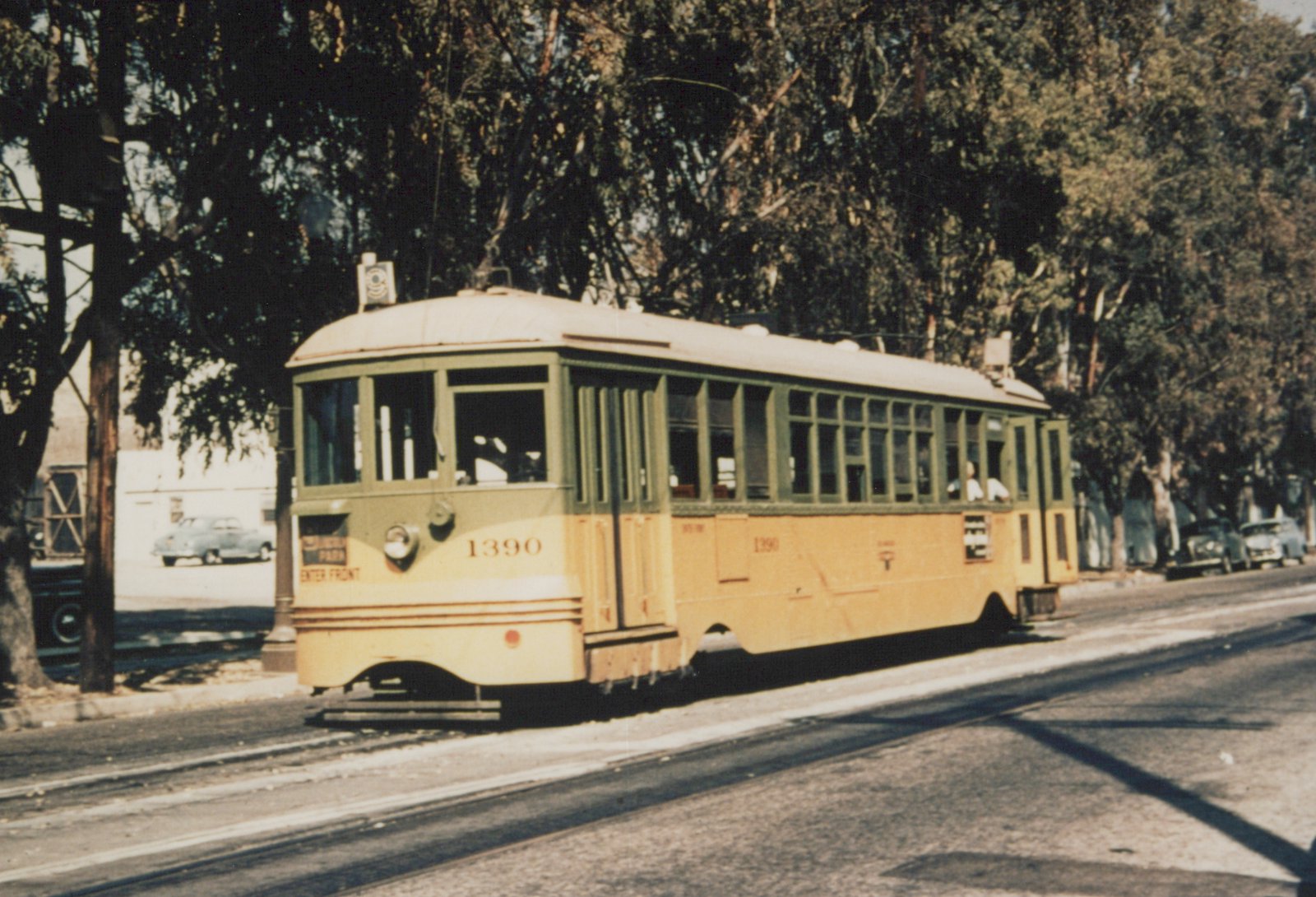 A yellow and green double-truck streetcar numbered 1390 on double street track under tall trees, with parked late-1940s cars in the background.