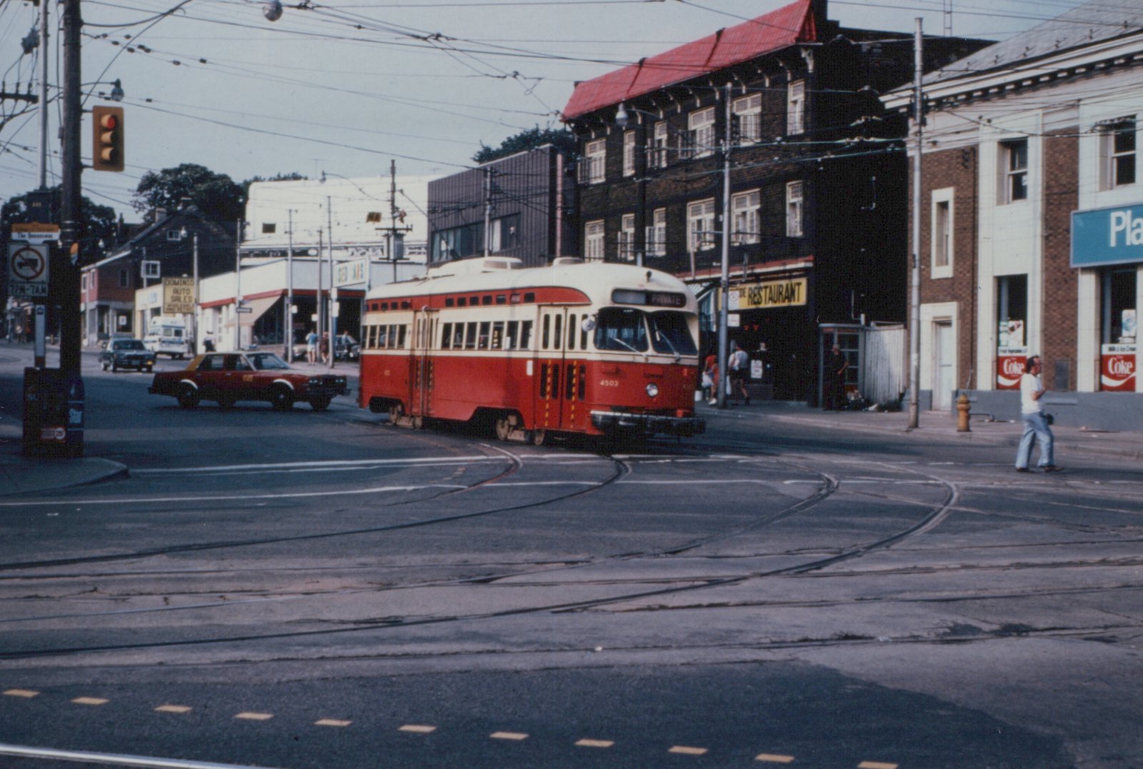 A red and cream streetcar numbered 4503 turns across a wide intersection laced with curving streetcar tracks under overhead wires, with brick buildings, a restaurant and a corner store behind it.