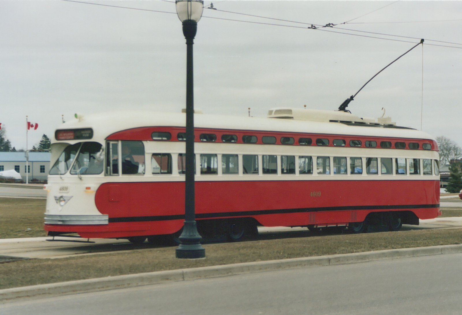 A red and cream streetcar numbered 4609 stands on a grassy track beside a street under overhead wire, with a lamp post in front of it and a Canadian flag in the distance.