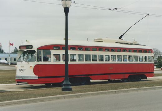 A red and cream streetcar numbered 4609 stands on a grassy track beside a street under overhead wire, with a lamp post in front of it and a Canadian flag in the distance.