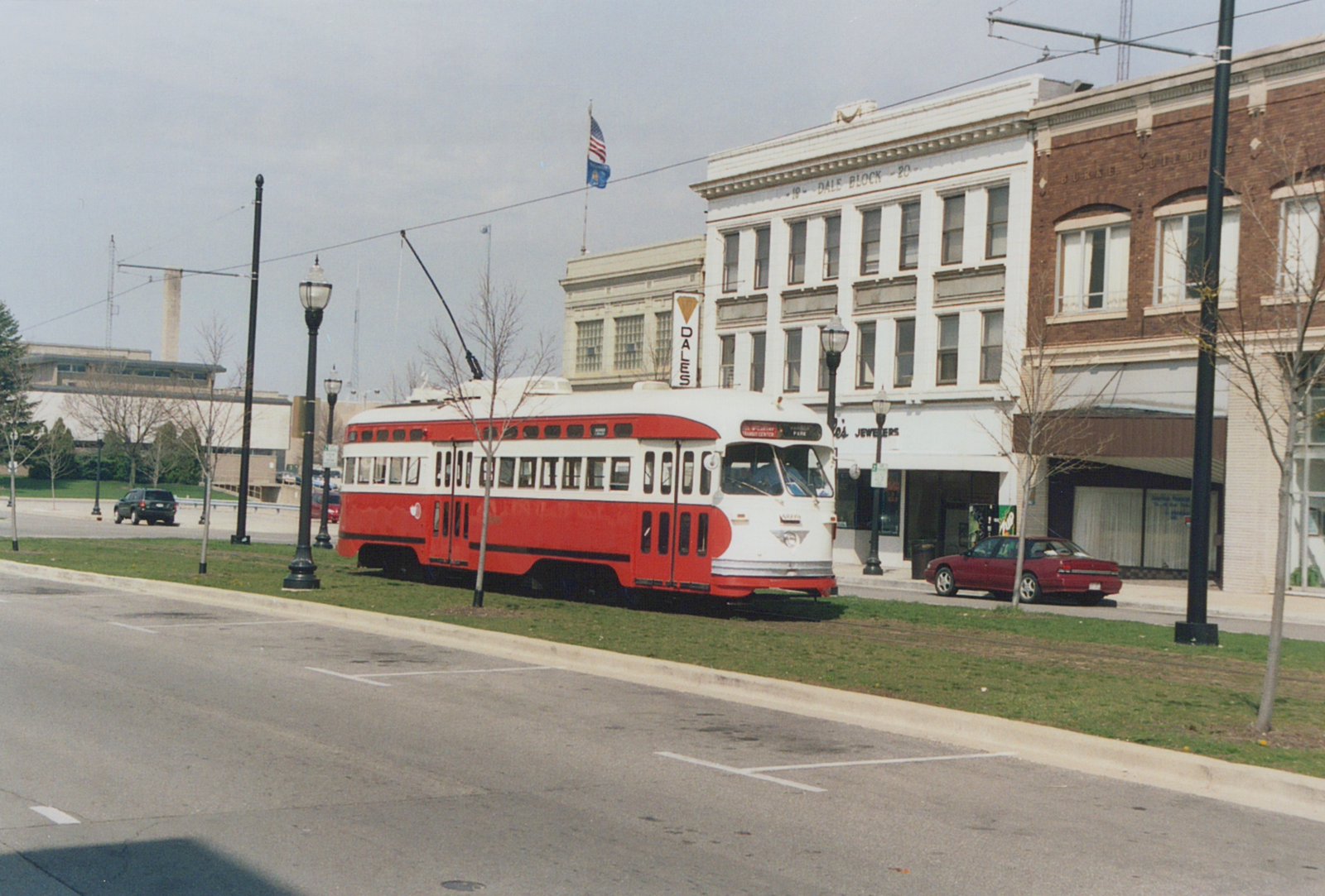 A red and white streamlined streetcar with its trolley pole raised runs on grass-covered track in a street median, in front of a white three-story commercial building and a red brick building, under an overcast sky.