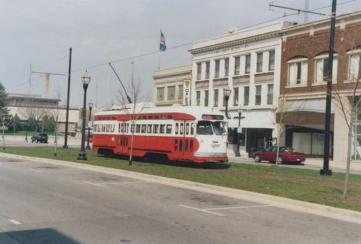 A red and white streamlined streetcar with its trolley pole raised runs on grass-covered track in a street median, in front of a white three-story commercial building and a red brick building, under an overcast sky.