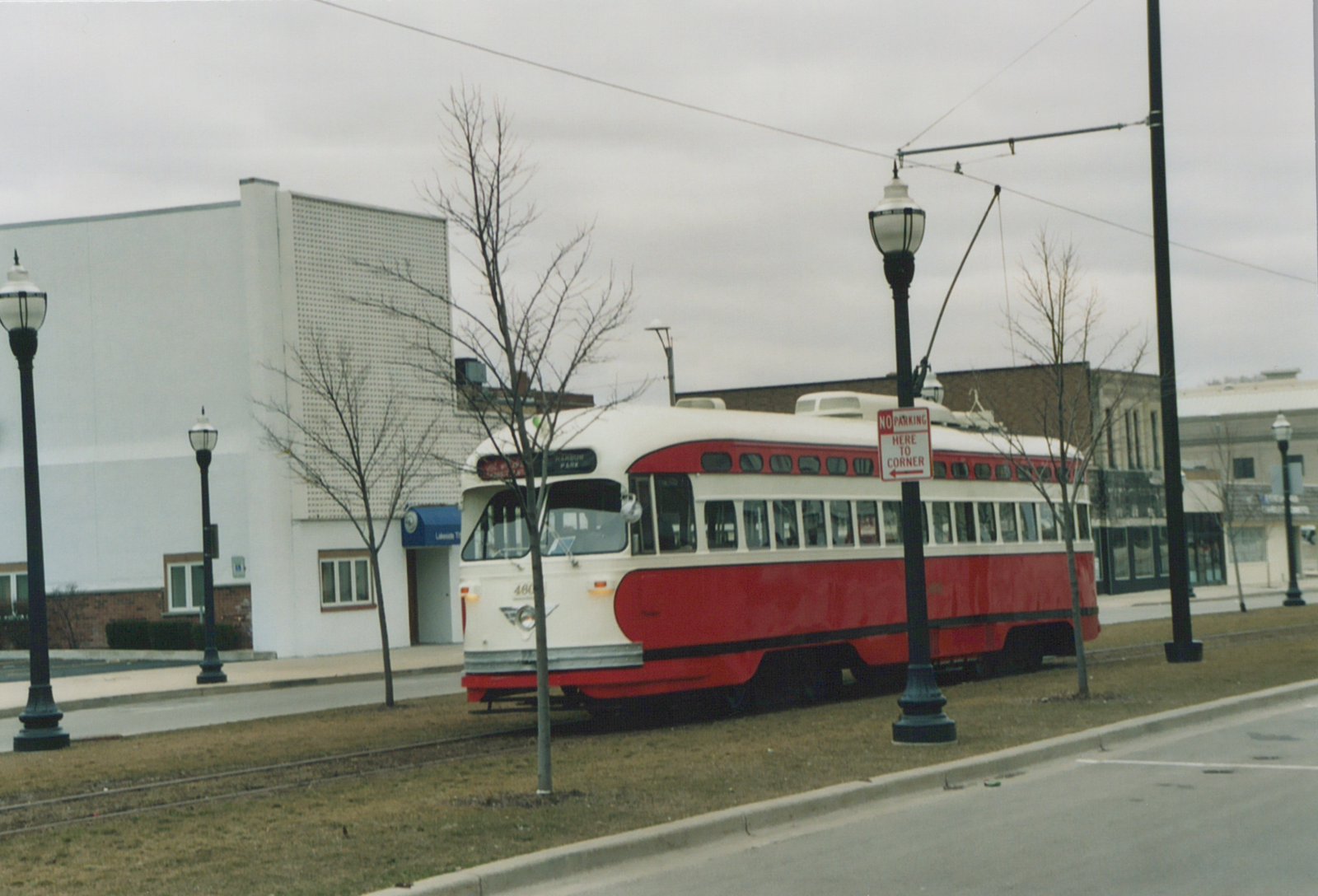 A red and cream streamlined streetcar with a trolley pole up sits on a single track in a grass median beside a street, with bare trees, ornamental lamp posts and a white building behind it under an overcast sky.
