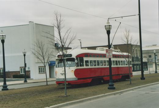 A red and cream streamlined streetcar with a trolley pole up sits on a single track in a grass median beside a street, with bare trees, ornamental lamp posts and a white building behind it under an overcast sky.